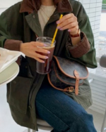 Une femme assise sur une chaise haute et accoudée à une table avec un verre dans une main et la paille jaune dans l'autre. Elle porte une veste mi saison verte avec les manches marrons en style velours