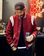 Blouson bomber homme à manches côtelées, style baseball, de couleur rouge, porté par un homme dans un bar, avec une casquette et un verre à la main