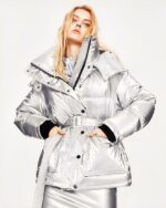Blouson chaud femme, mi-long, à capuche, col haut, avec ceinture pour définir la taille, de couleur gris argenté, porté par une femme blonde, avec les mains dans les poches