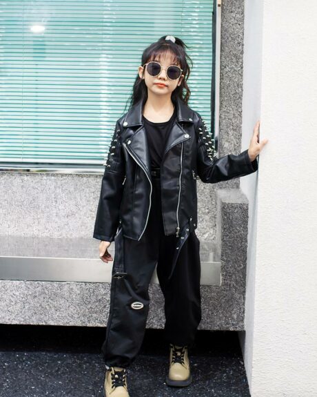 Une petite fille qui porte un Blouson de moto noir à clous devant une devanture de magasin.