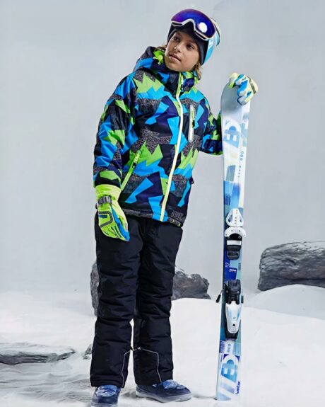 Un garçon en tenue de skir debout avec des skis dans la main.