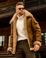 Un homme debout qui porte un blouson fourré marron en simili cuir avec un col rabattu fourré. Derrière lui se trouve un bâtiment marron et noir