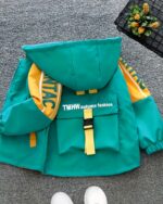 Blouson vert et jaune à capuche pour garçon posé au sol. Il a une poche dorsale. Le sol est gris.