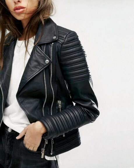 Une femme qui porte un blouson en simili cuir noir avec fermeture éclair. Elle porte un teeshirt blanc et elle a une main dans la poche de son pantalon