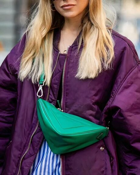 Une jeune femme blonde qui porte un blouson bombardier style grosse doudoune chaude couleur violette avec un sac banane vert