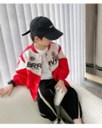 Garçon assis sur un canapé blanc portant un pantalon de jogging noir avec 2 rayures blanches. Un t-shirt blanc et un blouson de moto rouge et blanc avec des inscriptions et des logos sur la partie blanche.Il porte une casquette noire, une montre sur son poignet droit et un gobelet blanc dans sa main gauche. Derrière lui des stores styles vénitiens blancs.