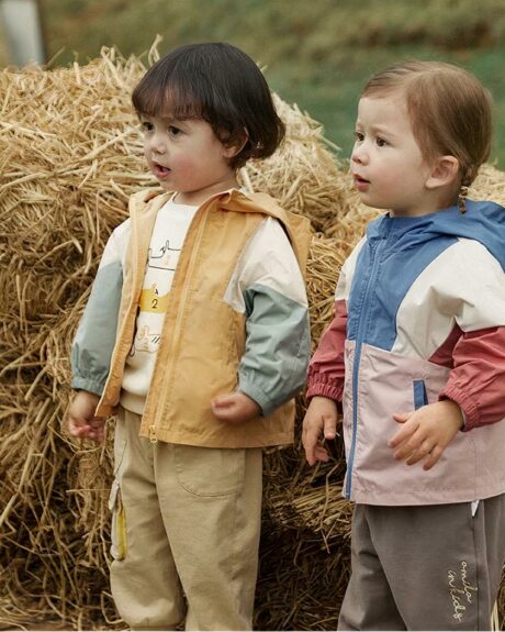 2 jeunes enfants debouts à côté d'une botte de paille, portant un blouson coloré à capuche.