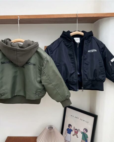 Deux blousons style aviateur à capuche effet pull. A gauche couleur vert militaire vue de dos, avec une inscription noir dans le dos. A droite, de face, un noir avec inscription blanche côté poitrine. Ils sont sur des cintres en bois. Au-dessus, il y a une étagère en bois. En dessous, un cadre avec contour noir avec écrit "we are family" et un abat-jour beige.