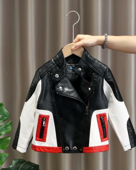 Blouson de moto en simili cuir pourenfant sur un cintre en bois tenu par la main d'une femme à droite. Il est noir sur le col et le buste, du blanc sur les manches et 2 poches devant avec une fermeture noire et encadrée de rouge. Le bord en bas est rouge. Double fermeture éclair devant avec un col tombant vers la gauche et la droite.