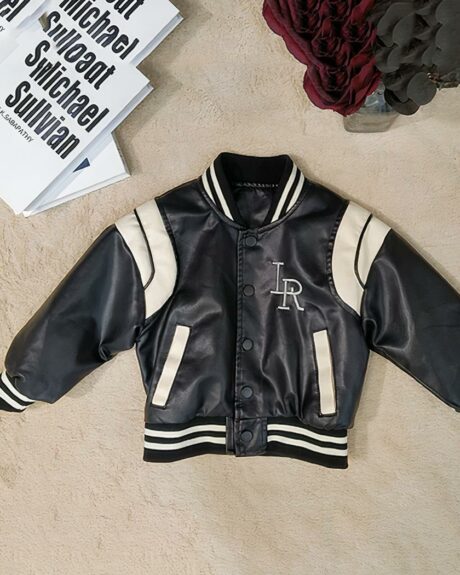 Blouson teddy pour garçon en simili cuir noir et blanc. Le blanc se retrouve au niveau des épaules et des poches latérales. Des rayures blanches sur le col, l'extrémité des manches et le bas du blouson. Les lettres L et R côté cœur. Le blouson est à pressions. Il est posé sur une moquette beige, des livres blancs en haut à gauche et des fleurs en haut à droite.