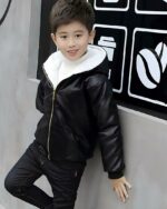 Jeune garçon type asiatique portant un pantalon noir et un blouson en simili cuir noir avec capuche et fermeture éclair. L'intérieur est fourré en blanc. Il est adossé à un mur gris avec une pancarte noire où il y a des symboles de grains de café et de gobelet.