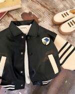 Blouson en simili cuir noir avec les manches blanches, 3 traits noirs sur la manche gauche. Avec pression et un motif de bip bip et le coyote sur le devant côté cœur. Un haut blanc est posé à l'intérieur du blouson. Le tout est posé sur un rond en bois. des chaussures beiges à droite, des livres en haut à gauche. Une main tient le bas du blouson.
