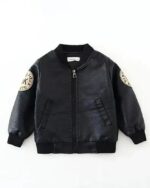 Blouson large en simili cuir noir pour garçon avec 2 poches latérales sans rabat et une fermeture éclair. Un motif beige en cercle sur chaque manche. Sur fond blanc.