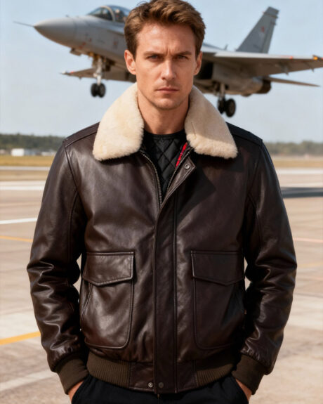 Un homme caucasien au regard très sérieux, portant un blouson aviateur homme en cuir marron avec col en fourrure couleur crème amovible devant un avion de chasse qui décolle.