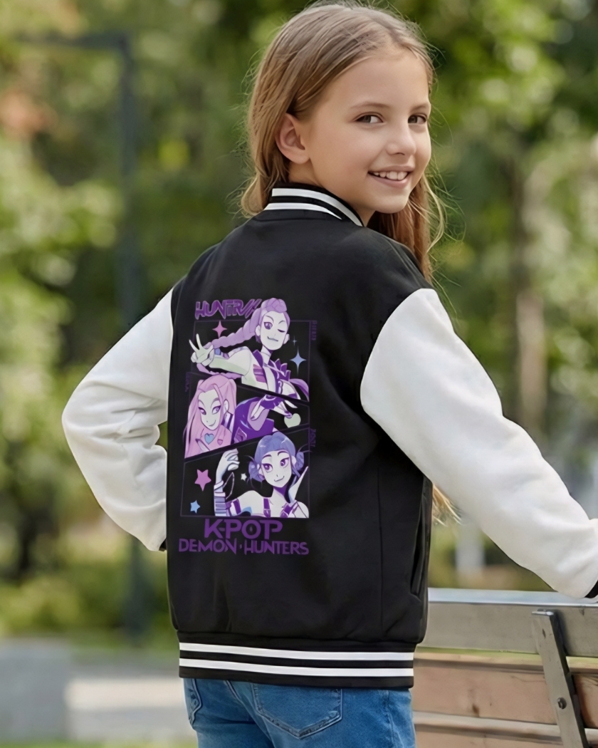 Une jeune fille souriante portant un blouson teddy enfant Kpop Demon Hunters Huntrix noir avec des manches blanches et un dessin violet au dos, posant près d'un banc en bois dans un parc verdoyant et ensoleillé.